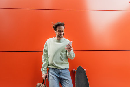 cheerful man in sweatshirt holding backpack and using smartphone near longboard and orange wall