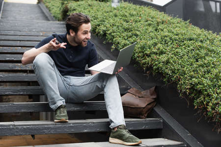 bearded man smiling and gesturing during video call on laptop while sitting on stairsの写真素材