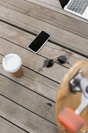 high angle view of gadgets with blank screen, sunglasses and paper cup on wooden table near blurred longboardの写真素材