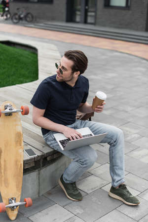 man in sunglasses and polo shirt holding paper cup and using laptop near smartphone and longboardの写真素材