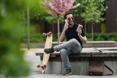 happy man in sunglasses holding paper cup and talking on smartphone near longboard and laptopの写真素材