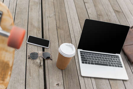 gadgets with blank screen, sunglasses and paper cup on wooden table near blurred longboardの写真素材