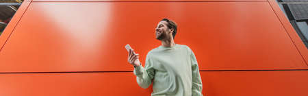 low angle view of cheerful man in sweatshirt holding smartphone near orange wall, bannerの写真素材