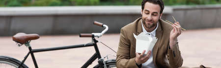 happy businessman in suit holding cardboard box with thai food and chopsticks while sitting near bicycle, bannerの写真素材