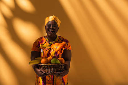 smiling middle aged african american woman in bright dress and turban holding basket with exotic...