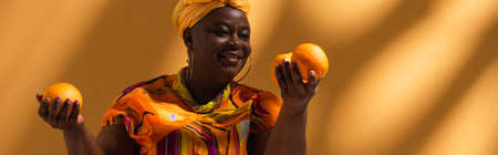 smiling middle aged african american woman holding oranges on background with shadows, bannerの写真素材