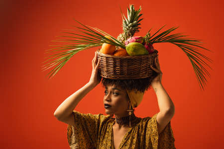 young african american woman holding basket with exotic fruits on head and looking away on redの写真素材