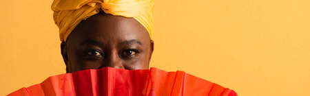 portrait of middle aged african american woman covering face with red fan on yellow, bannerの写真素材