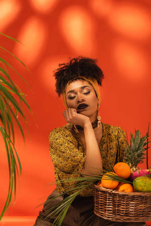 young african american woman with closed eyes and hand near face sitting near basket with exotic...