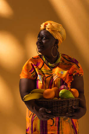 smiling middle aged african american woman holding basket with exotic fruits on orangeの写真素材