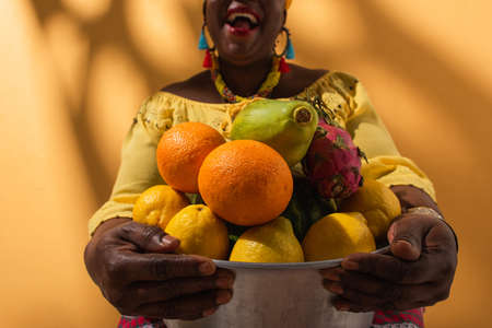 partial view of middle aged african american woman holding metal bowl with fruits on orangeの写真素材