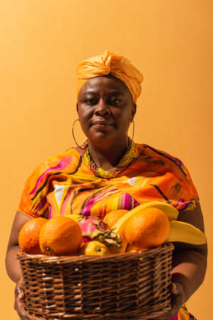 middle aged african american woman in bright dress holding basket with exotic fruits on orangeの写真素材