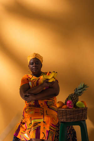 middle aged african american woman sitting near fruits and holding bananas on orangeの写真素材