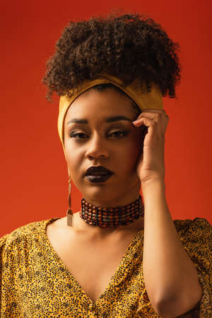 young african american woman with lipstick looking at camera with hand near face on red