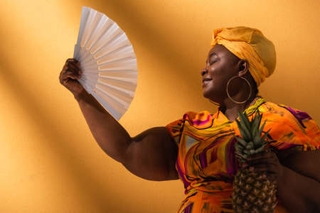 middle aged african american woman sitting with closed eyes, holding pineapple and waving with fan...