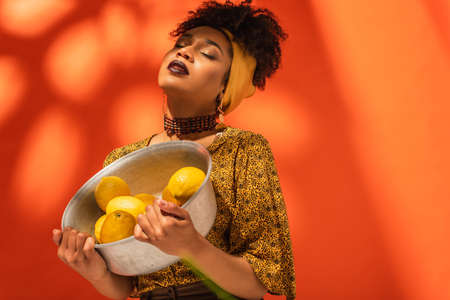 african american woman in blouse and headscarf holding metal bowl with lemons on orange