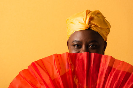 portrait of middle aged african american woman covering face with red fan on yellow