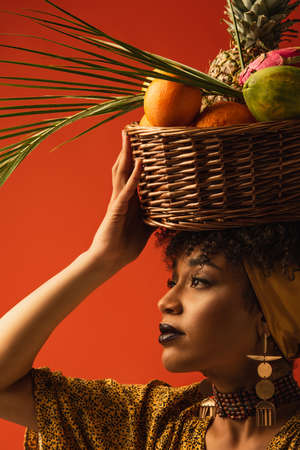 portrait of young african american woman holding basket with exotic fruits on head and looking away on redの写真素材