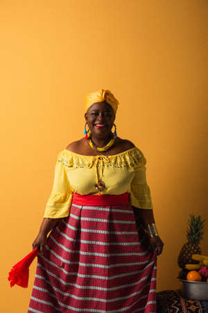middle aged african american woman in bright clothes and turban standing near fruits in metal bowl on yellowの写真素材