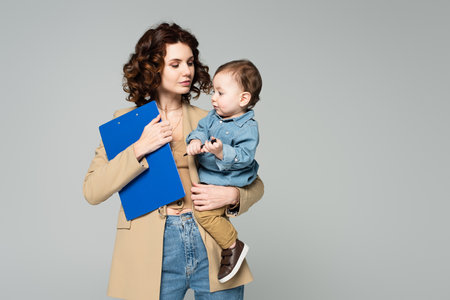 curly businesswoman in blazer holding clipboard and toddler boy in arms isolated on grayの写真素材