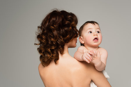 curly woman with bare shoulders holding in arms baby boy isolated on grayの写真素材