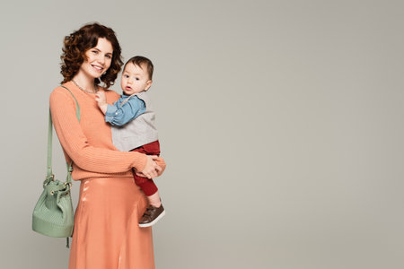 happy woman in satin skirt holding in arms toddler son isolated on gray