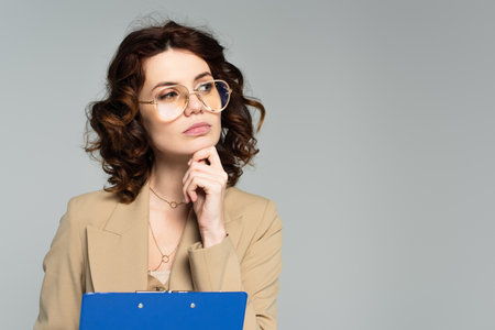 pensive young businesswoman in glasses and blazer holding clipboard isolated on grayの写真素材
