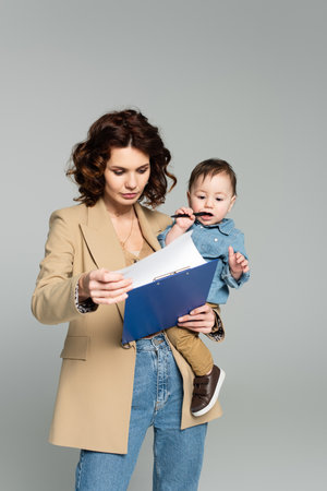 curly businesswoman in blazer holding clipboard and baby boy in arms isolated on grayの写真素材