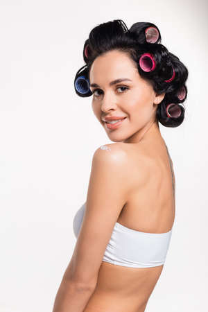 smiling young woman in curlers looking at camera with applied cream on shoulder isolated on whiteの写真素材