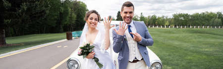Cheerful groom pointing at ring near bride with bouquet and vintage car, bannerの写真素材