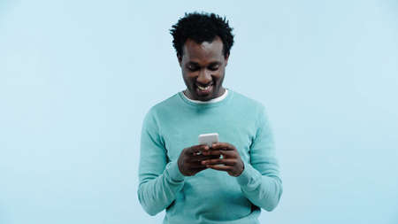 smiling african american man messaging on cellphone isolated on blueの写真素材