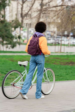 Back view of african american woman with backpack holding bicycle and walking outdoors