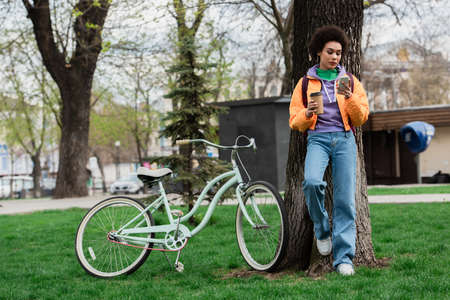 African american woman holding coffee to go and using smartphone near tree and bike outdoorsの写真素材