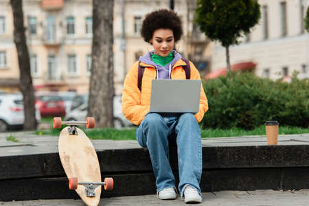 African american freelancer using laptop near coffee to go and longboard outdoorsの写真素材