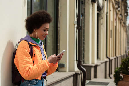 Smiling african american woman with backpack using smartphone near building
