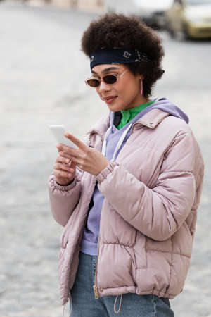 African american woman in bandana and sunglasses using smartphone outdoors
