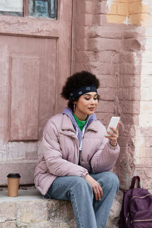 Stylish african american woman using smartphone near paper cup and backpack on stair of building