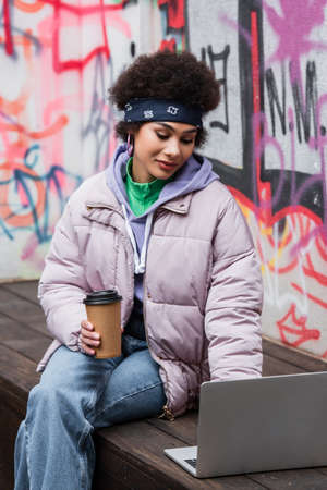 Stylish african american freelancer with paper cup using laptop on bench near graffiti outdoors