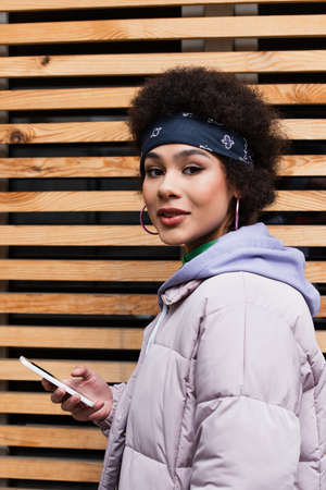 African american woman in jacket holding mobile phone and looking at camera outdoorsの写真素材