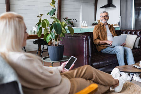 Smiling elderly man using laptop near blurred wife with cellphone in living roomの写真素材