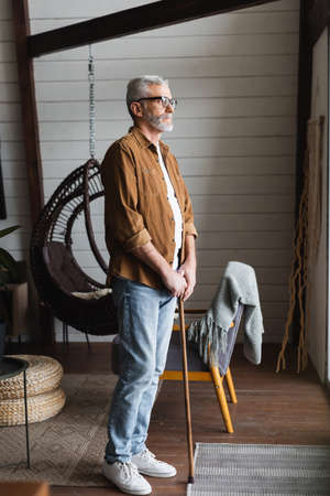 Senior man in eyeglasses holding crutch while standing at homeの写真素材