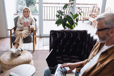 Senior woman using smartphone near blurred husband with laptop on couchの写真素材
