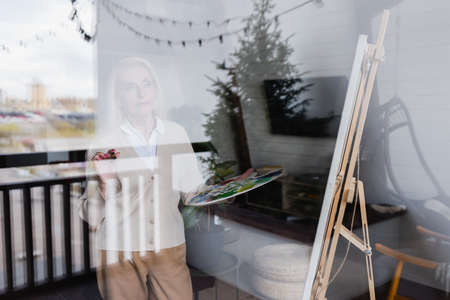 Senior woman holding palette near easel and window at homeの写真素材