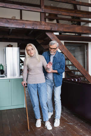 Senior woman with crutch standing near careful husband at homeの写真素材