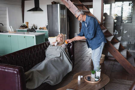Man holding pills and glass of water near ill wife on couchの写真素材