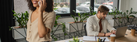young man working with laptop near african american colleague talking on cellphone in modern office,...