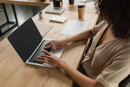 partial view of young african american woman sitting at desk with paper cup and typing on laptop in modern officeの写真素材