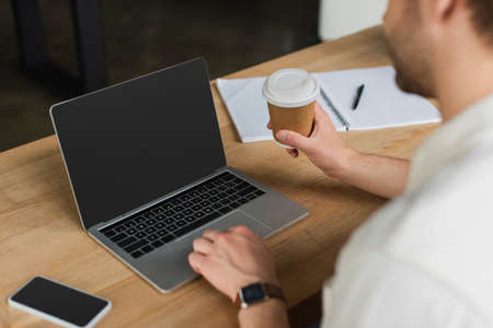 partial view of young man sitting with paper cup in hand and typing on laptop with blank screen in modern officeの写真素材