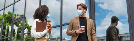 young interracial colleagues in medical masks walking with paper cups and documents near glass...