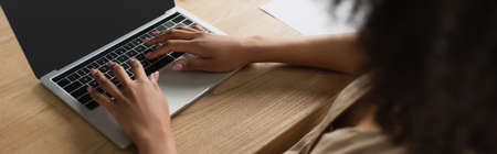 partial view of young african american woman sitting at desk and typing on laptop in modern office, bannerの写真素材